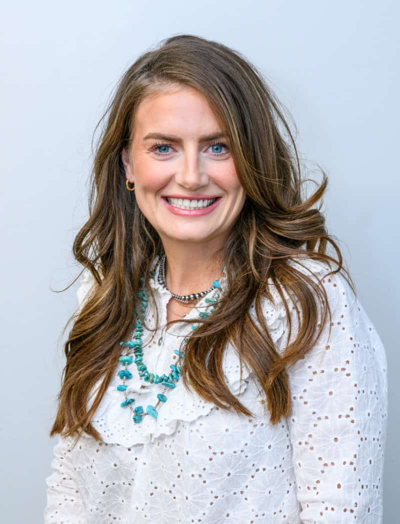 High-quality woman smiling with long brown hair wearing white eyelet blouse and turquoise jewelry.